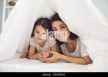 Felice giovane mummia e carina piccola figlia che guarda la macchina fotografica nascondendosi sotto il morbido copriletto bianco Foto Stock