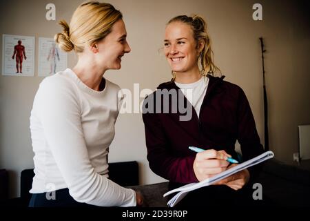 Giovane paziente femminile e fisioterapista sorridente mentre indica la documentazione seduta sulla curva di massaggio in studio. Foto Stock