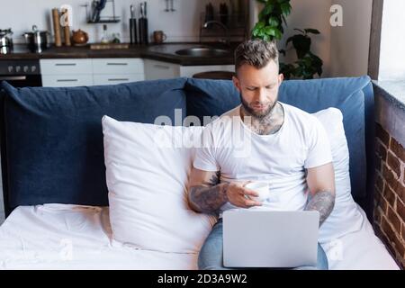 giovane freelance tatuato che usa il computer portatile a letto mentre sorregge una tazza di caffè Foto Stock