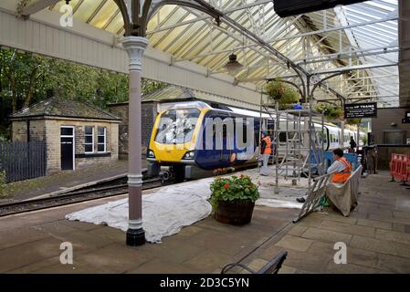 Pittori e decoratori che lavorano sulla piattaforma come un treno di Classe 196 arriva alla stazione ferroviaria di Hebden Bridge, Yorkshire, Regno Unito Foto Stock