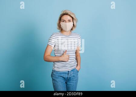 Bella signora bionda con maschera medica sul viso è gesturing il segno simile su una parete blu dello studio Foto Stock