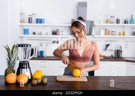 Giovane casalinga che taglia l'arancia vicino alla frutta e il frullatore sulla cucina tabella Foto Stock