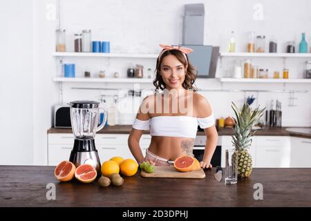 Giovane donna che guarda la macchina fotografica vicino alla frutta e al frullatore tabella Foto Stock