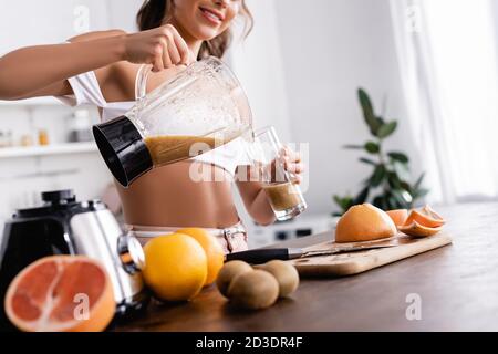 Vista tagliata di giovane donna che versa frullato in vetro vicino frutta biologica sul tavolo da cucina Foto Stock