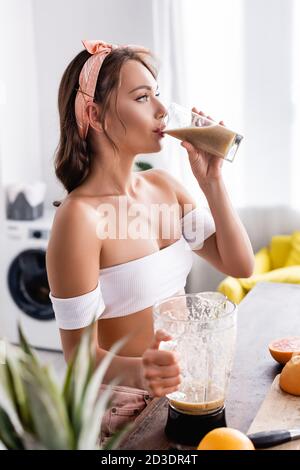 Fuoco selettivo di frullato giovane donna che beve vicino al frullatore e. frutta a casa Foto Stock