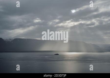 Una nave da crociera soluna in un fiordo e cielo drammatici paesaggio con alberi di luce solare che brillano attraverso le nuvole Dell'Islanda settentrionale Foto Stock