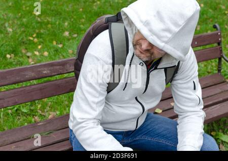 Un uomo di mezza età in un maglione con cappuccio seduto da solo una panca di parcheggio Foto Stock