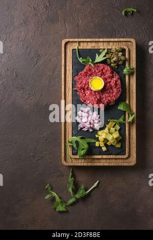 Tartare di manzo con uovo di quaglia in guscio, taglio cetrioli sottaceto, capperi, cipolla rossa, erba cipollina, rucola servita su legno nero ardesia board con pepe mil Foto Stock