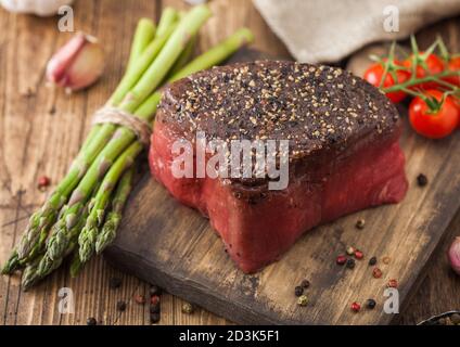 Fetta di manzo crudo fesa giunto con il sale e il pepe sul tagliere di legno con pomodori aglio e punte di asparagi su legno tavolo da cucina. Foto Stock