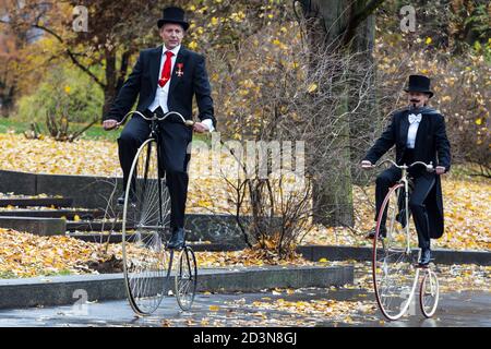 Due ciclisti su biciclette Penny Farthing che indossano un giro sul cappotto di coda attraverso il parco autunnale Foto Stock