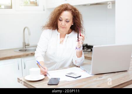 Donna anziana si è concentrata sulla revisione di carte e fatture. È nella cucina della sua casa. Foto Stock