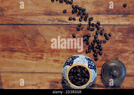 Vasetto in ceramica con motivo blu e bianco su piano di legno, riempito con chicchi di caffè crudo. Coperchio spento, i chicchi di caffè fuoriuscito attraverso il tavolo. Vista dall'alto. Foto Stock