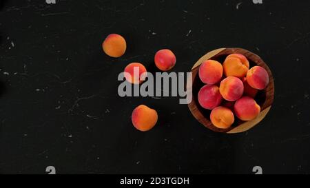 Albicocche mature in piccola ciotola di legno, alcune sparse su marmo nero come scrivania accanto ad essa. Vista dall'alto, spazio per il testo a sinistra Foto Stock