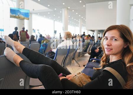 Giovane donna seduta nella sala dell'aeroporto, stanca dopo aver aspettato un paio d'ore per il volo di collegamento in prima mattina, le gambe sul sedile di fronte a h. Foto Stock