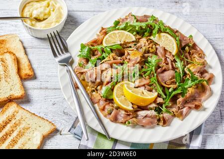 insalata di tonno a fette sottili con capperi, rucola, pinoli e salsa di senape su un piatto bianco su un tavolo di legno con toast alla griglia e burro morbido, h Foto Stock