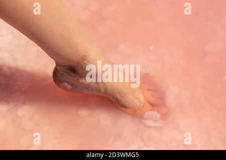 Donna gamba piede primo piano in rosa fiocchi di sale vista dall'alto sotto la superficie d'acqua rosa. Procedure termali sane Foto Stock