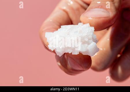 Dita delle mani che tengono rosa bianco fiocco di sale formazione di cristallo vicino rosa vibrante superficie d'acqua. Centro benessere soleggiato primo piano Foto Stock