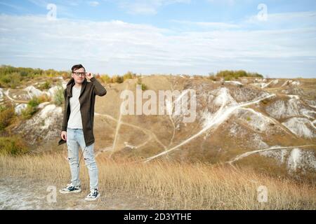 Uomo con occhiali in jeans, t-shirt e cape si erge in terreno collinare. Il maschio adulto nel cappuccio regola i suoi occhiali e gode di una bella vista di Foto Stock