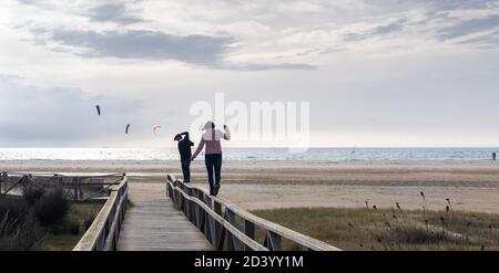 Madre e figlio che si divertono a camminare in equilibrio su un Recinzione nella spiaggia di Tarifa Valdevaqueros con kitesurf sullo sfondo Foto Stock