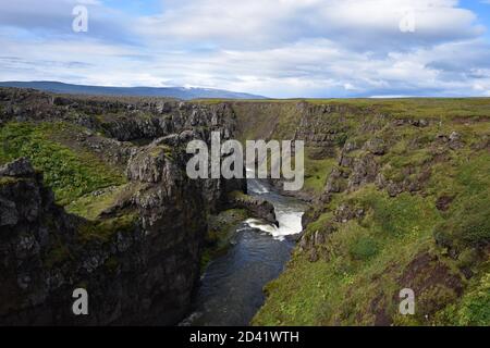 Il fiume Víðidalsá scorre attraverso il Canyon di Kolugljufur nel nord dell'Islanda. Muschio verde e erba linee la scogliera. Montagne visibili in lontananza. Foto Stock
