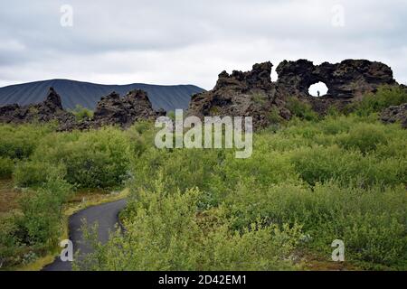 Il campo di lava a Dimmuborgir nel Lago Myvatn, Islanda del Nord. Un sentiero pedonale attraverso gli alberi. Una finestra di lava con un visitatore all'interno. Hverfell dietro. Foto Stock