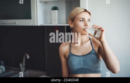 Bella donna caucasica con i capelli biondi che indossano gli sportswear sta bevendo un bicchiere d'acqua durante le lezioni di fitness digitale presso casa Foto Stock