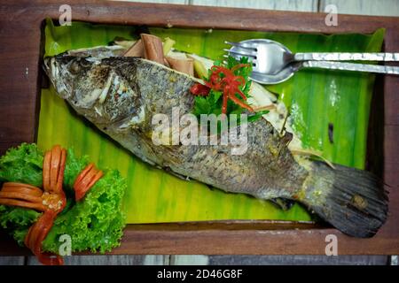 Snapper bianco alla griglia intero e farcito con erbe arrangiate su foglie di banana in un vassoio di legno splendidamente decorato con verdure e paprika, tailandese Foto Stock