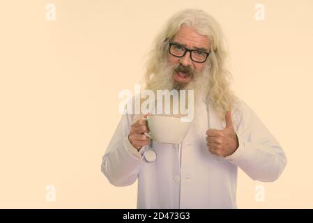 Studio shot di felice senior uomo barbuto medico sorridere mentre dando pollice in alto e tenendo premuto tazza da caffè Foto Stock