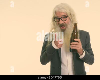 Studio shot di felice senior imprenditore barbuto sorridere mentre si tiene una bottiglia di birra e dando pollice in alto Foto Stock