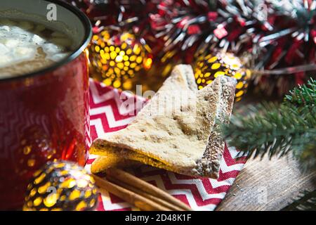 Biscotti triangolari con zucchero in polvere e cannella su un tovagliolo con motivo a zigzag nell'arredamento natalizio. Tazza di caffè rosso con marshmallow, abete rosso Foto Stock