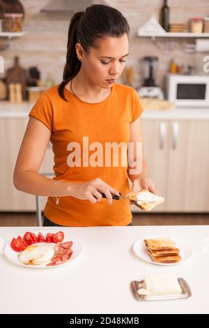 Preparazione del panino per la prima colazione con burro spalmato sul pane tostato. Donna che stende il burro sul pane tostato per colazione. Coltello spalmato burro morbido su fetta di pane. Uno stile di vita sano, che prepara un pasto delizioso al mattino in una cucina accogliente. Pranzo tradizionale gustoso Foto Stock