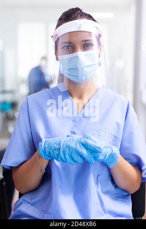 Assistente medico con visiera e maschera facciale stanchi in sala d'attesa dell'ospedale durante l'epidemia di coronavirus. Medico che consulta l'uomo anziano in sala esame. Medico, epidemia, cura, chirurgia, corridoio. Foto Stock