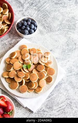 Minuscoli pancake per colazione. Frittelle di cereali con mirtilli, banane, fragole su sfondo grigio. Cibo alla moda. Spazio di copia per testo o disegno Foto Stock