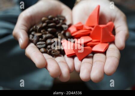 una mano da donna che tiene i chicchi di caffè e la carta origami rossa a forma di cuore. caffè arrosto con aroma fresco pronto per essere preparato Foto Stock