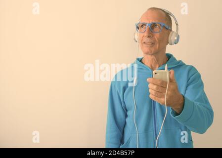 Studio shot di calvo uomo anziano che tiene il telefono cellulare mentre pensare e ascoltare musica con gli occhiali Foto Stock