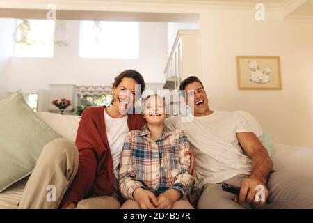 Sorridente famiglia seduto sul divano godendosi guardare la televisione a casa. Famiglia di tre persone che si rilassa a casa guardando la tv. Foto Stock