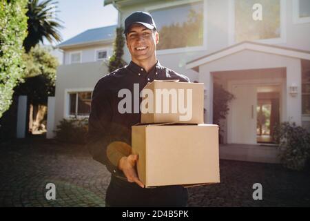 Corriere sorridente che consegna pacchi scatole. Persona di consegna maschile che tiene scatole di cartone all'aperto guardando la macchina fotografica e sorridendo. Foto Stock