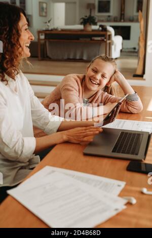 Madre sorridente che presta attenzione a sua figlia mentre lavora su un computer portatile a casa. Madre e figlia seduti insieme a casa con computer portatile e digitale Foto Stock