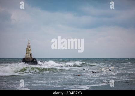 I surfisti di wave board in neoprene nero aspettano onde più grandi per catturare e navigare sul faro di Ahtopol, Bulgaria. Mar Nero. Onde di mare che spruzzi e freddo duro tempo ventoso Foto Stock