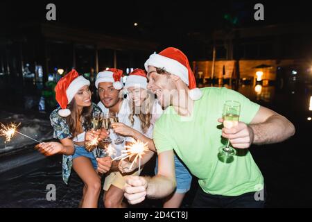 Fuoco selettivo degli amici con gli sparklers che si aggrappano con champagne vicino piscina di notte Foto Stock