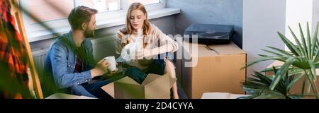 fuoco selettivo della donna gioiosa che tiene la tazza mentre disimballando la scatola con il boyfriend, colpo panoramico Foto Stock