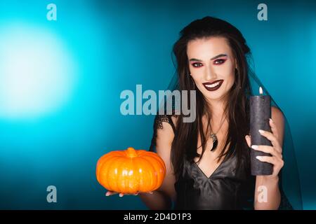 donna gioiosa con trucco nero e velo che tiene la candela bruciante e la zucca su blu Foto Stock