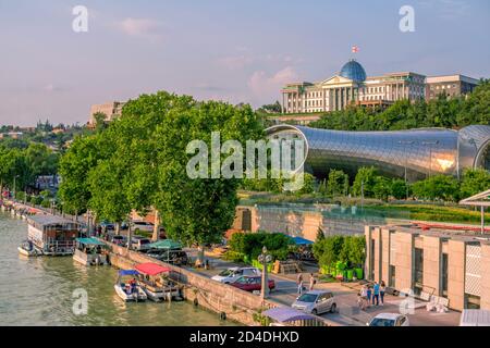 Tbilisi/Georgia - 19 luglio 2019: Paesaggio urbano estivo - fiume Kura dal ponte, palazzo del presidente e edificio del centro culturale, alberi verdi e blu di sk Foto Stock