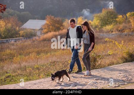 Una giovane e felice famiglia cammina con un cane Schnauzer lungo la strada, tenendo le mani, guardarsi l'un l'altro su uno sfondo di cielo blu e foresta. Foto Stock