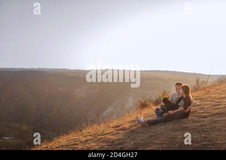 Seduto su un canyon, guarda i parapendio che volano nel cielo al tramonto. Viaggio con animali domestici. Giovane famiglia con un cane schnauzer viaggio. Moldavia, Vecchia Orhey Foto Stock