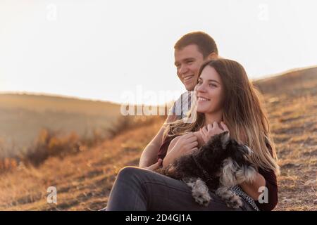 Giovane famiglia con un cane schnauzer viaggio. Bella ragazza abbraccia un giovane e tiene un animale domestico tra le braccia. Seduto su un canyon e guardando i parapendio Foto Stock