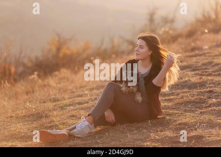 Bella ragazza giovane con lunghi capelli rossi che si sviluppano nel vento abbraccia un cane nero Schnauzer. Guarda la macchina fotografica e sorride al tramonto. Cura degli animali domestici Foto Stock