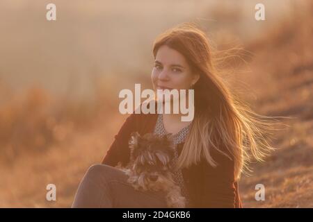 Felice giovane donna viaggia con il suo cane domestico nero schnauzer. Una bella ragazza abbraccia un animale domestico, si siede su una montagna, il vento sviluppa i suoi lunghi capelli rossi, Foto Stock