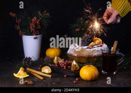 Torta fatta in casa ricoperta di zucchero in polvere e bacche rosse. Mano con sparkler bruciante. VIN brulé, rami di abete, coni di abete e mandarini su una b scura Foto Stock