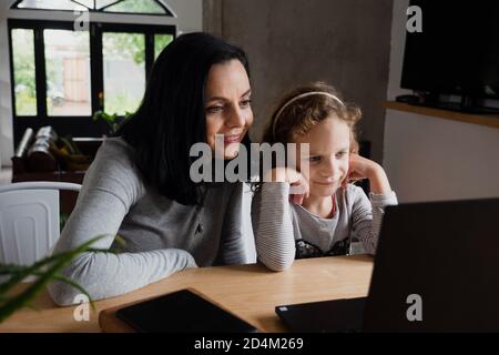 Madre felice e figlia del capretto che guardano il laptop, la mamma sorridente e la ragazza del bambino che si diverte con video in linea sul calcolatore Foto Stock